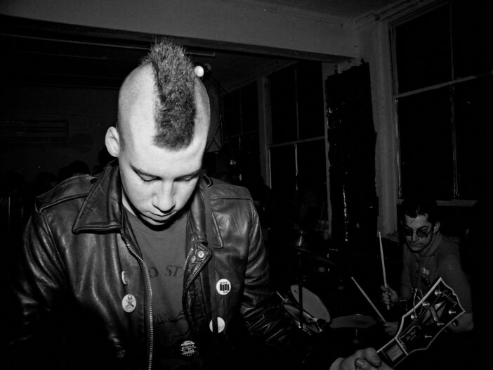 Punk musician in black and white playing in a small intimate venue, head down and focused, independent of any corporate machine.